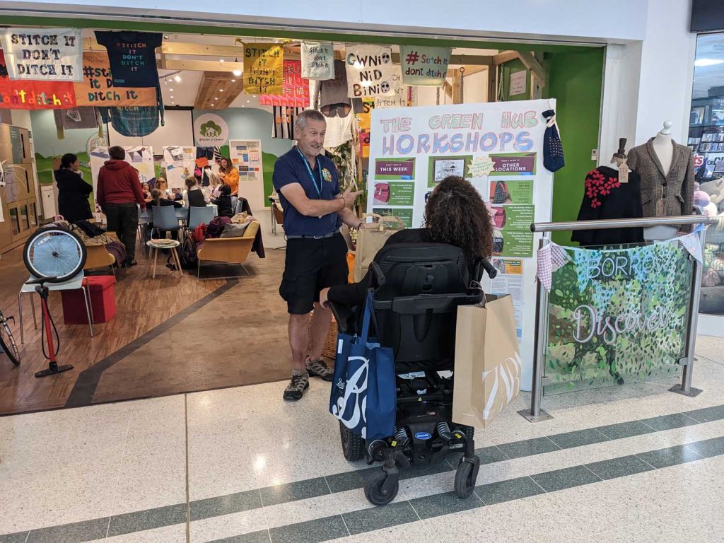 Photo of a person standing by a sign talking to a person in a wheelchair.
